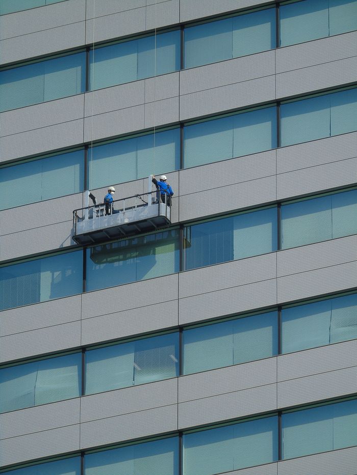 services-01 Two workers clean windows on a modern glass building facade using a suspended platform.