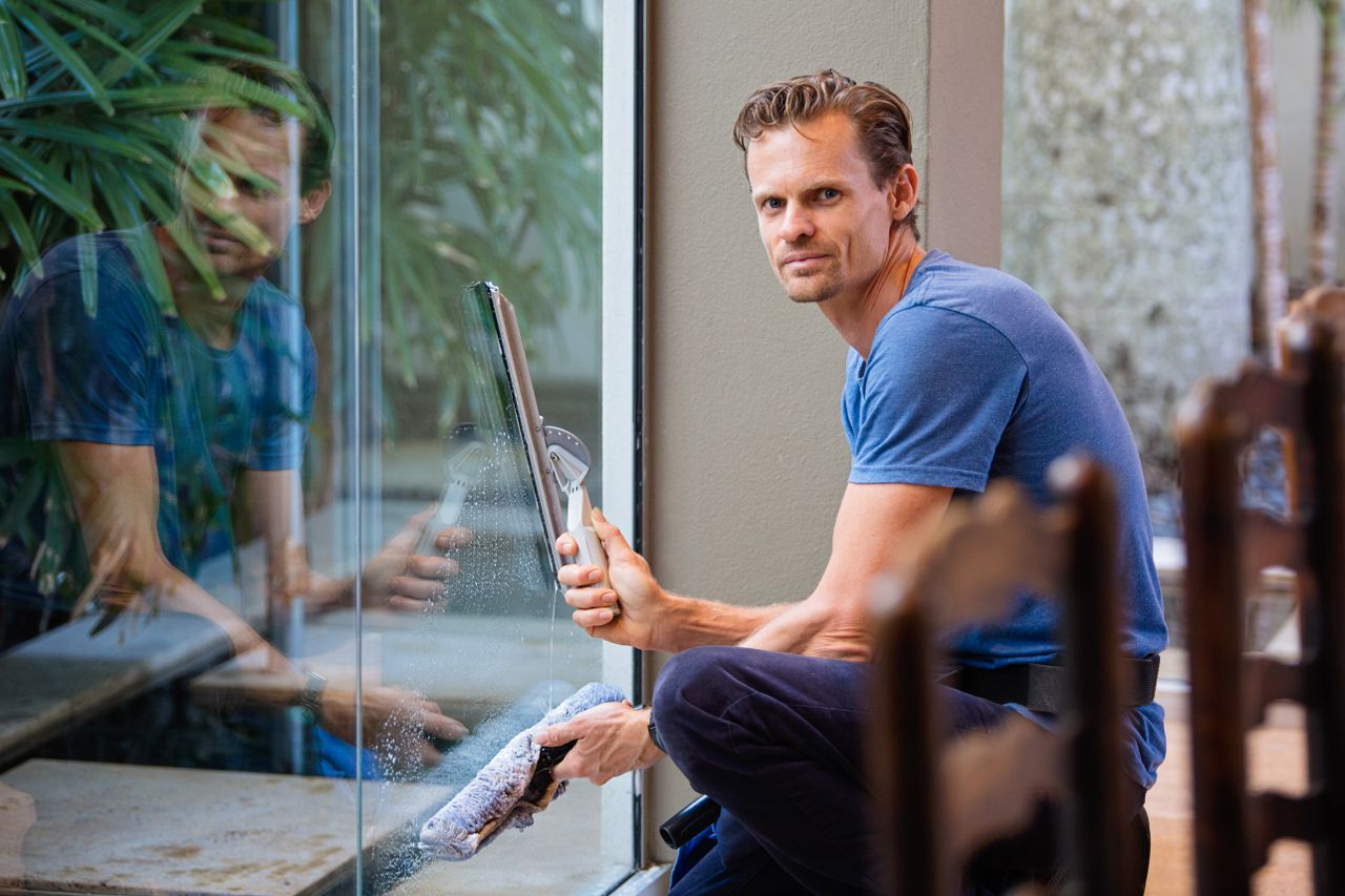our-story A man in a blue t-shirt cleaning large glass pane with a squeegee indoors.