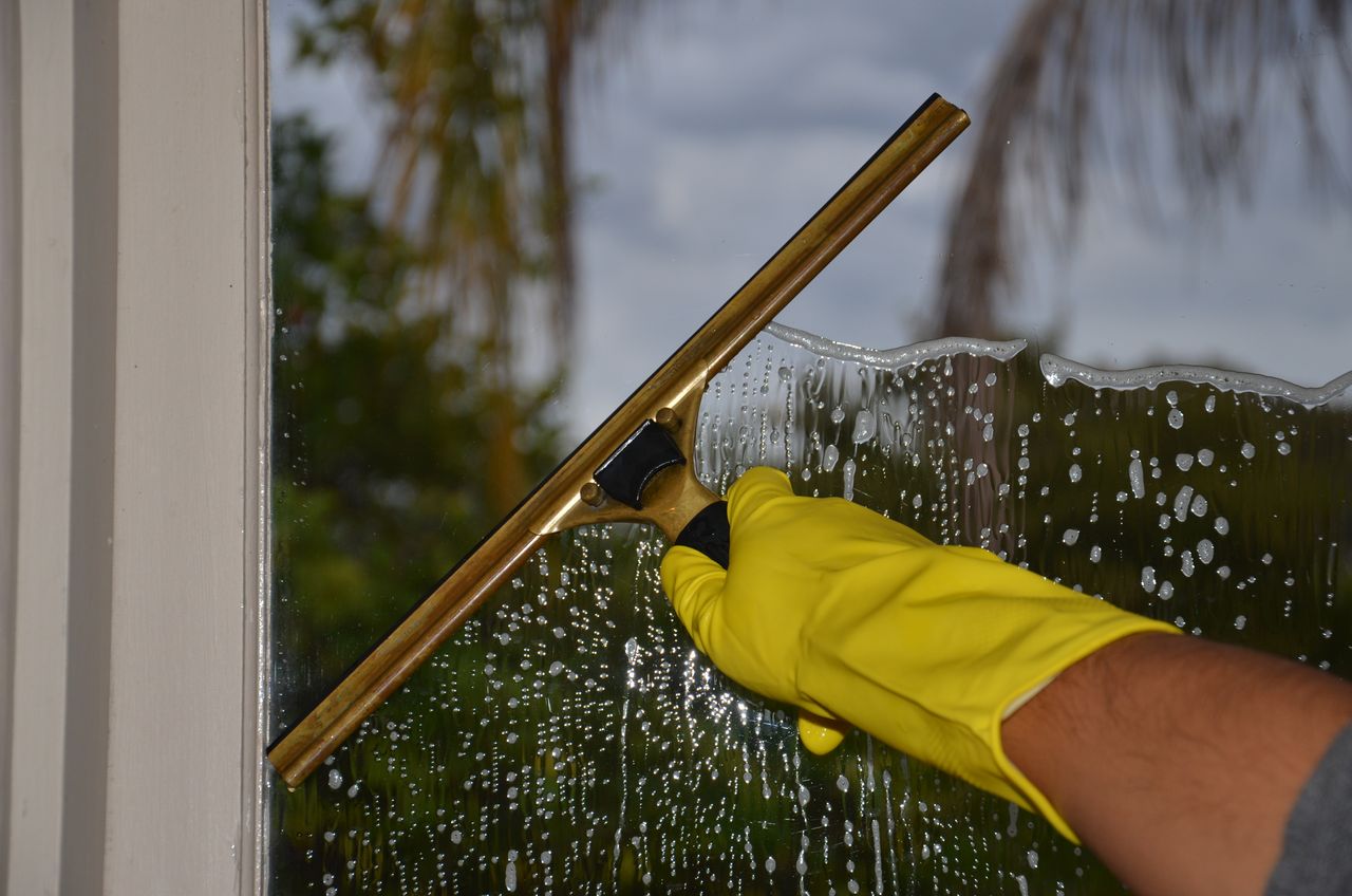 heros-img Close-up of hand cleaning window with squeegee outdoors using yellow gloves.